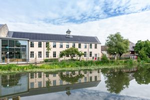 A slate-roof building with a pond and a bridge in front, surrounded by lush greenery.