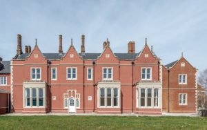 Front facade of a historic manor house painted terracotta red with white bay windows and multiple tall chimneys.