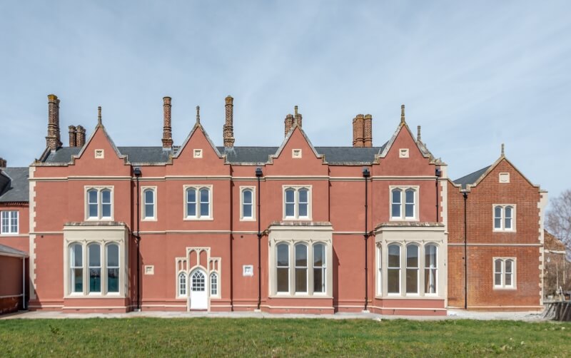 Front facade of a historic manor house painted terracotta red with white bay windows and multiple tall chimneys.