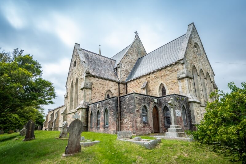 Wide-angle view of a historic stone church with pointed arch windows, slate roofing, and a surrounding graveyard with old headstones.