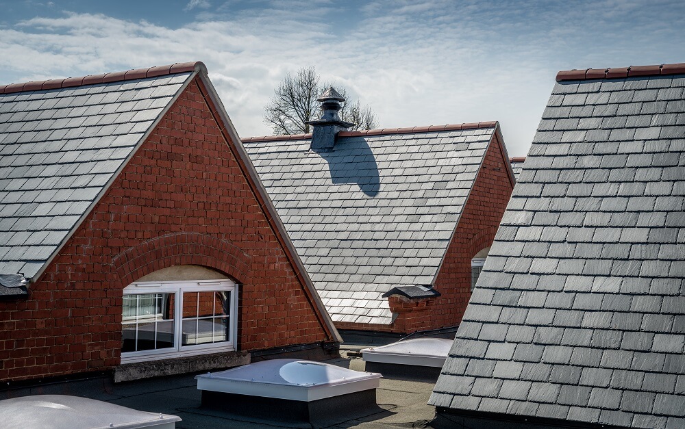 View of several pitched roofs covered with grey slate tiles on red-brick buildings, with a rooftop vent and skylights visible on the flat roof areas below