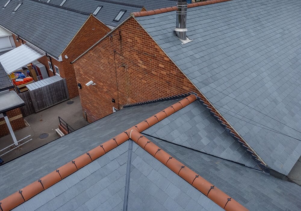 Aerial view of intersecting grey slate roofs on red-brick buildings, showing terracotta ridge tiles and a metal chimney, with a courtyard and outbuildings below