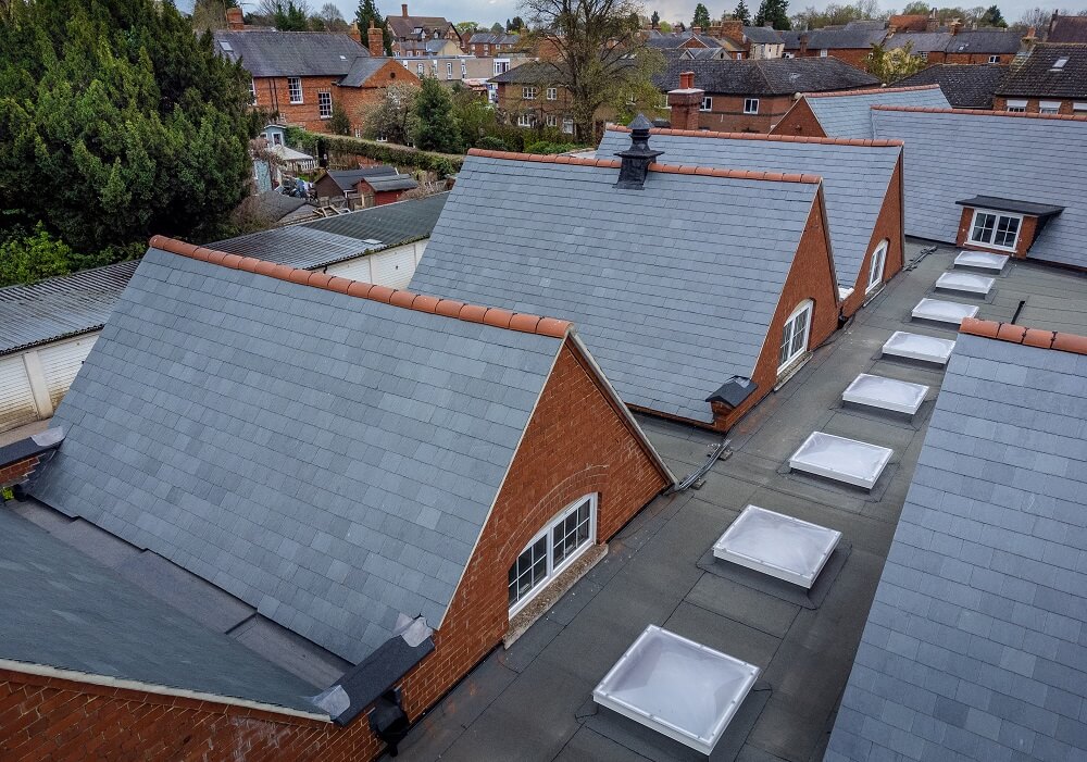 Elevated view of a row of red-brick buildings with grey slate pitched roofs, looking down onto multiple rectangular skylights installed on the flat roof corridor between them