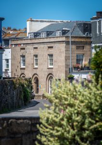 A wide shot of a historic two-story building made of light-colored stone blocks. The ground floor features arched doorways and windows, while the upper floor has rectangular sash windows. A blurred flowering bush is in the foreground.