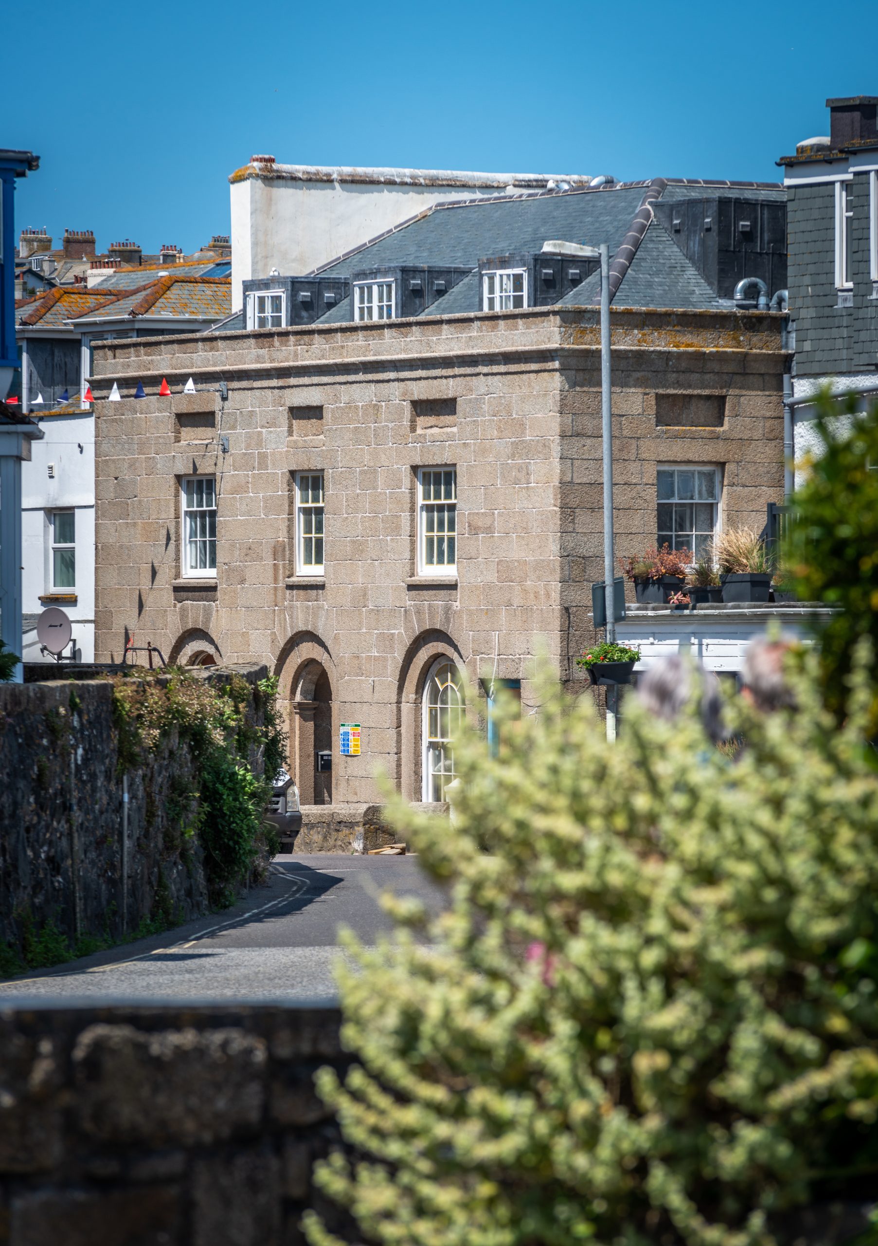 A wide shot of a historic two-story building made of light-colored stone blocks. The ground floor features arched doorways and windows, while the upper floor has rectangular sash windows. A blurred flowering bush is in the foreground.