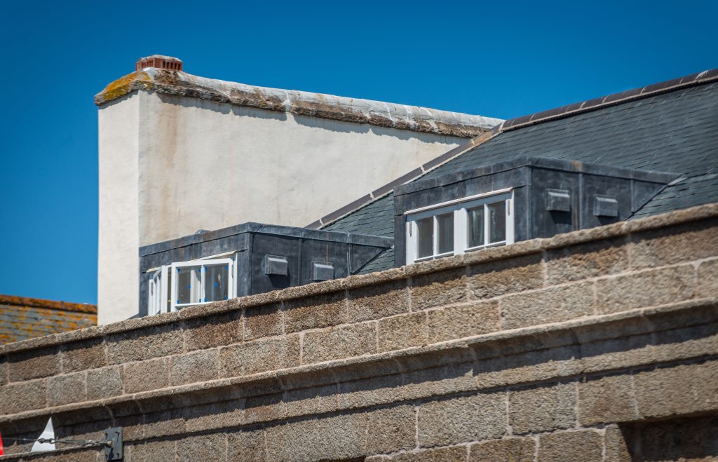 A close-up view of the top of the stone building, showing a grey slate mansard roof with two lead-clad dormer windows. A large, white-rendered chimney stack stands to the left against a clear blue sky.