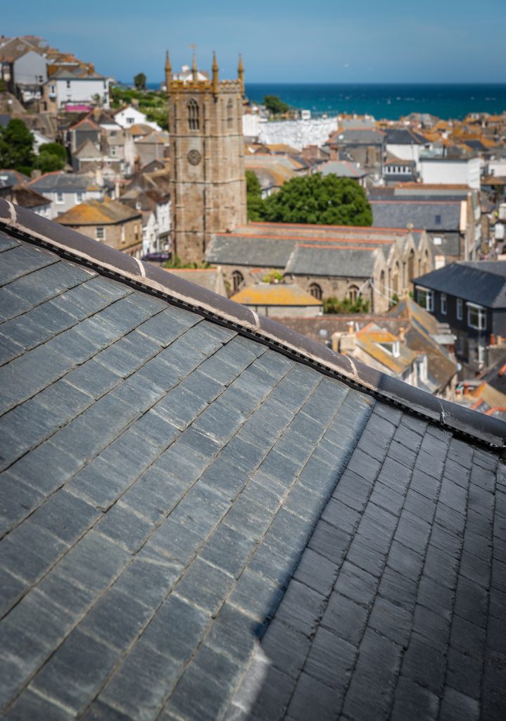 A high-angle view looking over a grey slate roof toward a coastal town. In the background, a historic stone church tower rises above the rooftops, with the blue ocean visible on the horizon.