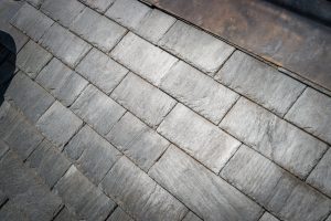 A detailed close-up of overlapping grey slate roof tiles. The texture of the natural stone is visible, showing various shades of charcoal and light grey under bright sunlight.