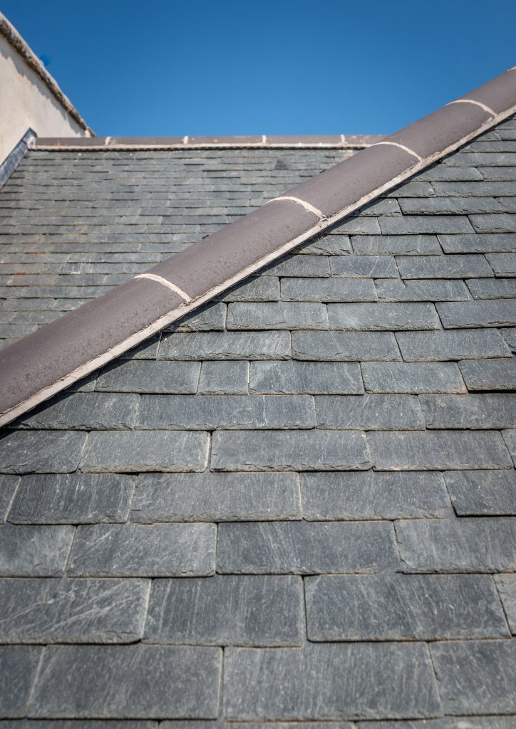 A view looking up a slate roof slope toward a rounded ridge cap. The slate tiles are neatly laid in rows, and the ridge is finished with dark, semicircular tiles and light-colored mortar.