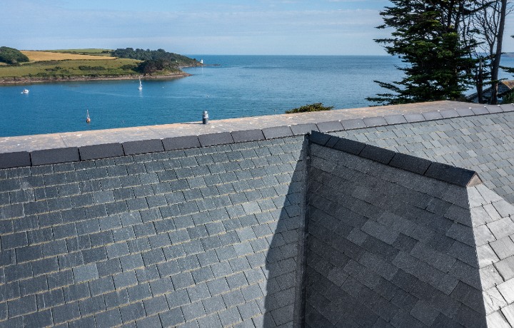 A high-angle view looking over the textured grey slate roof tiles toward a stunning blue bay with sailboats in St Mawes.