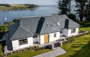 An elevated wide shot of the entire coastal residence, showing how the grey natural slate roof complements the white masonry and the surrounding ocean landscape.