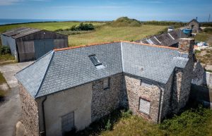 A wide shot showing the L-shaped stone cottage and slate roof against the backdrop of the exposed North Devon coastline and the Atlantic Ocean.