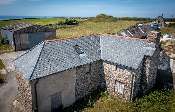 A wide shot showing the L-shaped stone cottage and slate roof against the backdrop of the exposed North Devon coastline and the Atlantic Ocean.