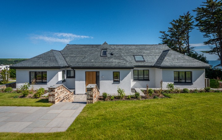 A front elevation of the completed home, showcasing the expansive natural slate roof, modern wooden door, and stone-walled entryway.