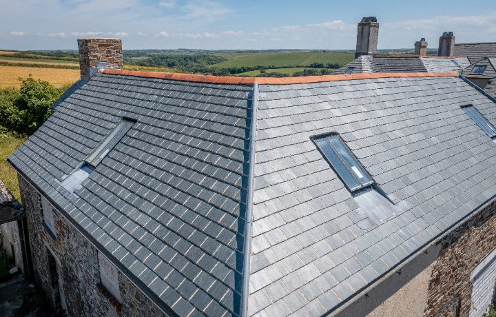 A top-down view of the roof meeting point, showing the precise installation of the slates and skylights overlooking the rolling Devon countryside.