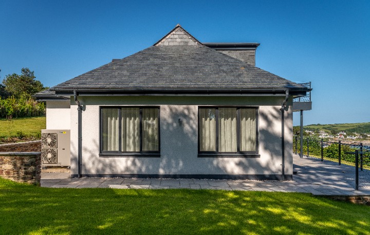 A side view of Tregenna featuring a crisp white exterior and a complex hipped roof clad in grey Riverstone Ultra Natural Slate, with a modern air source heat pump installed at the base.