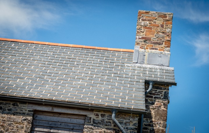 A detailed shot of the stone chimney stack integrated with the new slate roofing, featuring lead flashing and grey guttering.